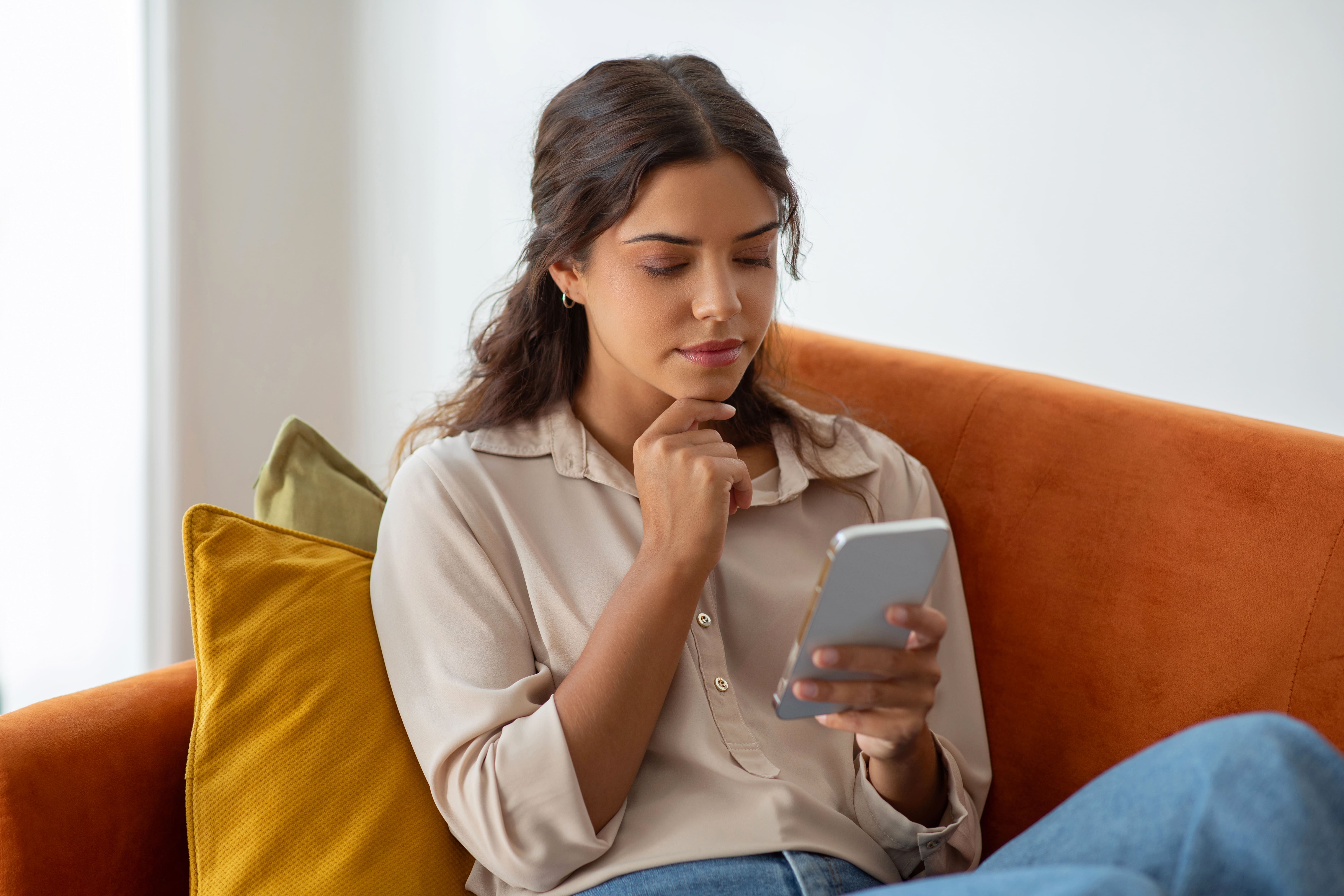 thoughtful-young-woman-touching-chin-while-reading-message-smartphone-home (1)
